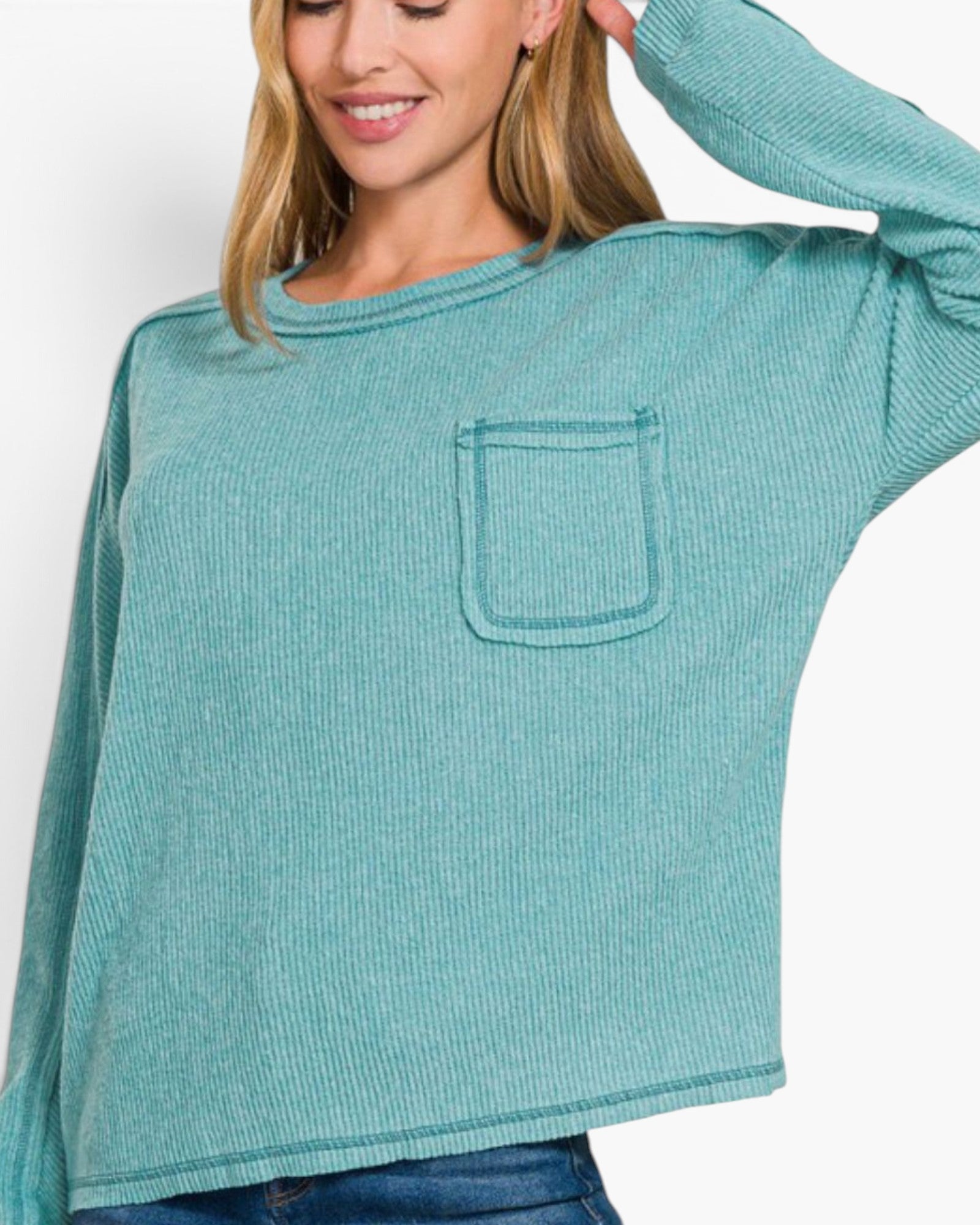 A woman wearing a teal ribbed long-sleeve top with a chest pocket, paired with blue jeans, against a plain white background.