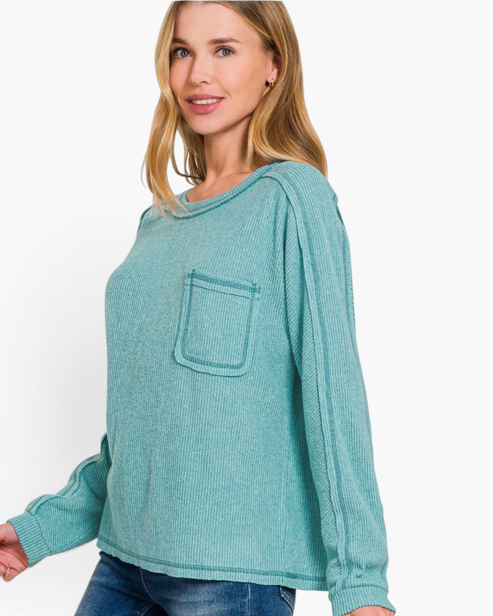 A woman wearing a teal long-sleeve ribbed knit top with a chest pocket, paired with blue jeans, against a plain white background.