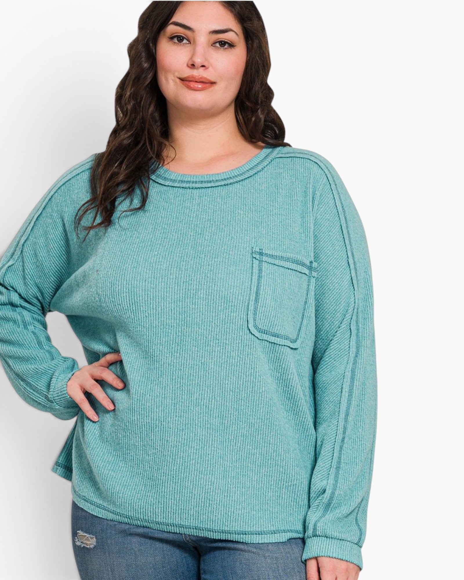 A woman wearing a ribbed knit long-sleeve top in a muted teal green color, featuring a chest pocket, paired with blue jeans against a plain white background.
