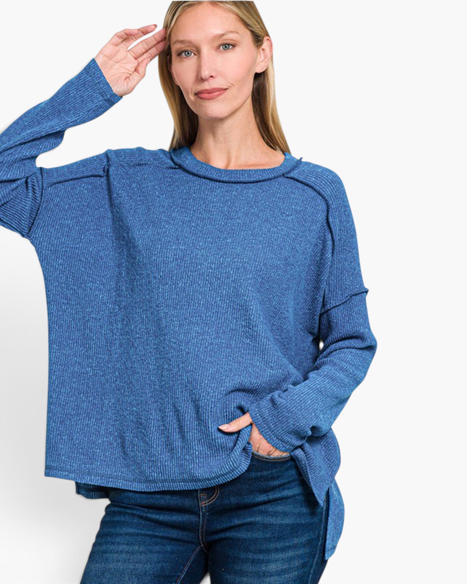 A woman wearing a heathered blue long-sleeve knit sweater and dark blue jeans, posing against a clean white studio background.