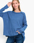 A woman wearing a heathered blue long-sleeve knit sweater and dark blue jeans, posing against a clean white studio background.