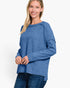 A woman wearing a heathered blue long-sleeve knit top with a wide boat neckline and drop shoulders, paired with dark blue jeans, against a plain white studio background.