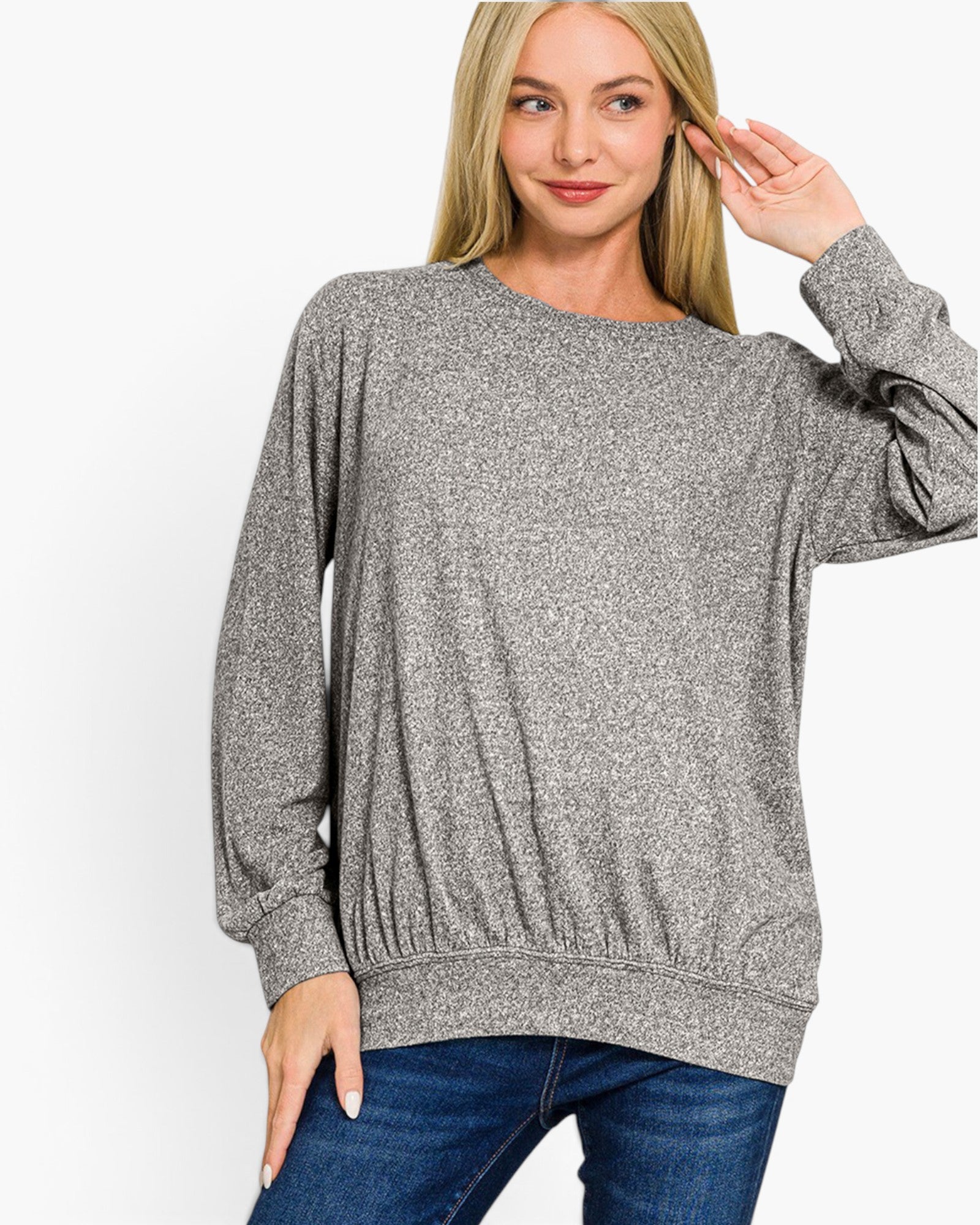 A woman wearing a heather gray long-sleeve crewneck sweater and blue jeans, posing against a solid white studio background.