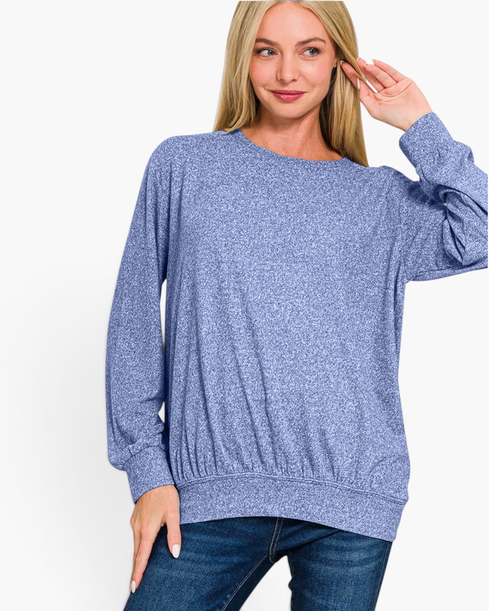 A woman wearing a heathered periwinkle blue long-sleeve crewneck sweater and dark blue jeans, posing against a solid white studio background.