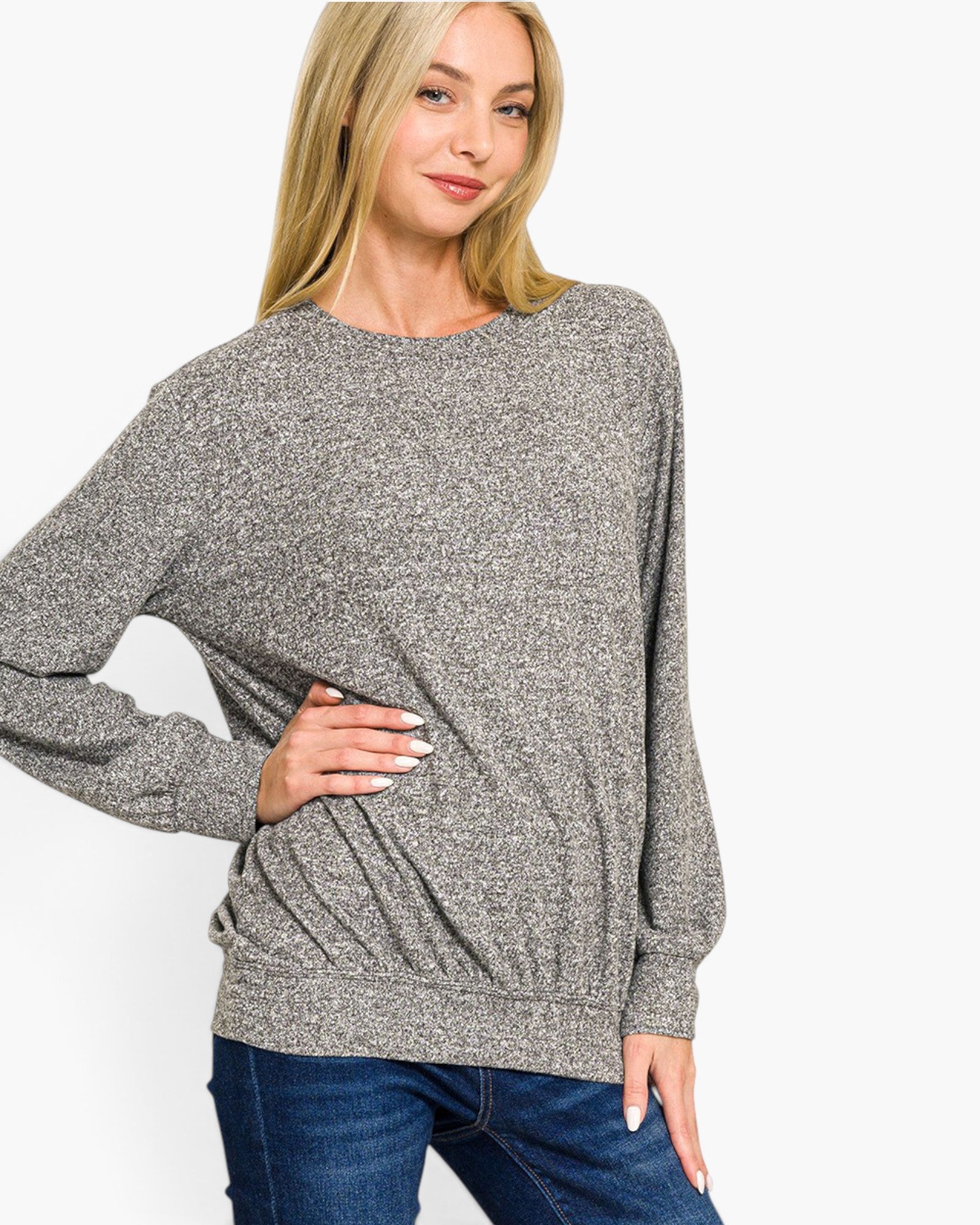 A woman wearing a heather gray long-sleeve crewneck sweater and blue jeans, posing against a solid white studio background.