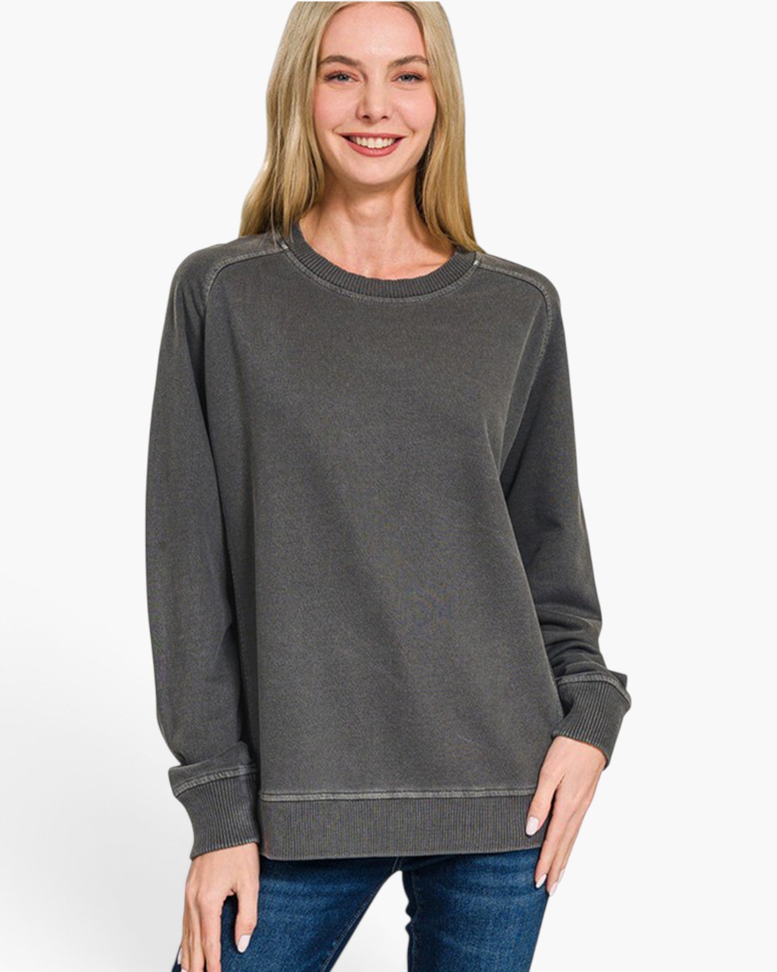 A woman wearing a dark gray crewneck sweatshirt and blue jeans, standing against a plain white studio background.