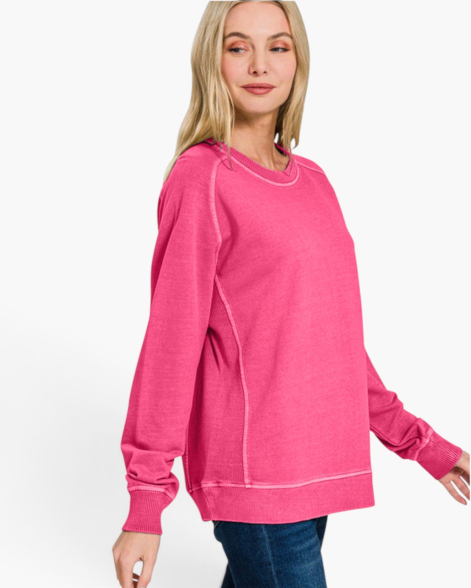 A woman wearing a bright pink crewneck sweatshirt with contrast white stitching, paired with blue jeans, photographed against a clean white studio background.