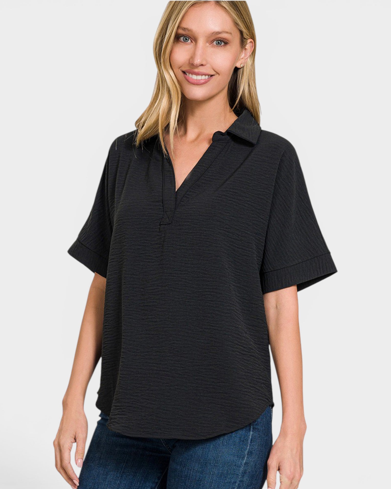 A woman wearing a black short-sleeve collared top with a textured, ribbed fabric and a partial button placket, paired with blue jeans, against a light gray background.