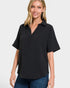 A woman wearing a black short-sleeve collared top with a textured, ribbed fabric and a partial button placket, paired with blue jeans, against a light gray background.