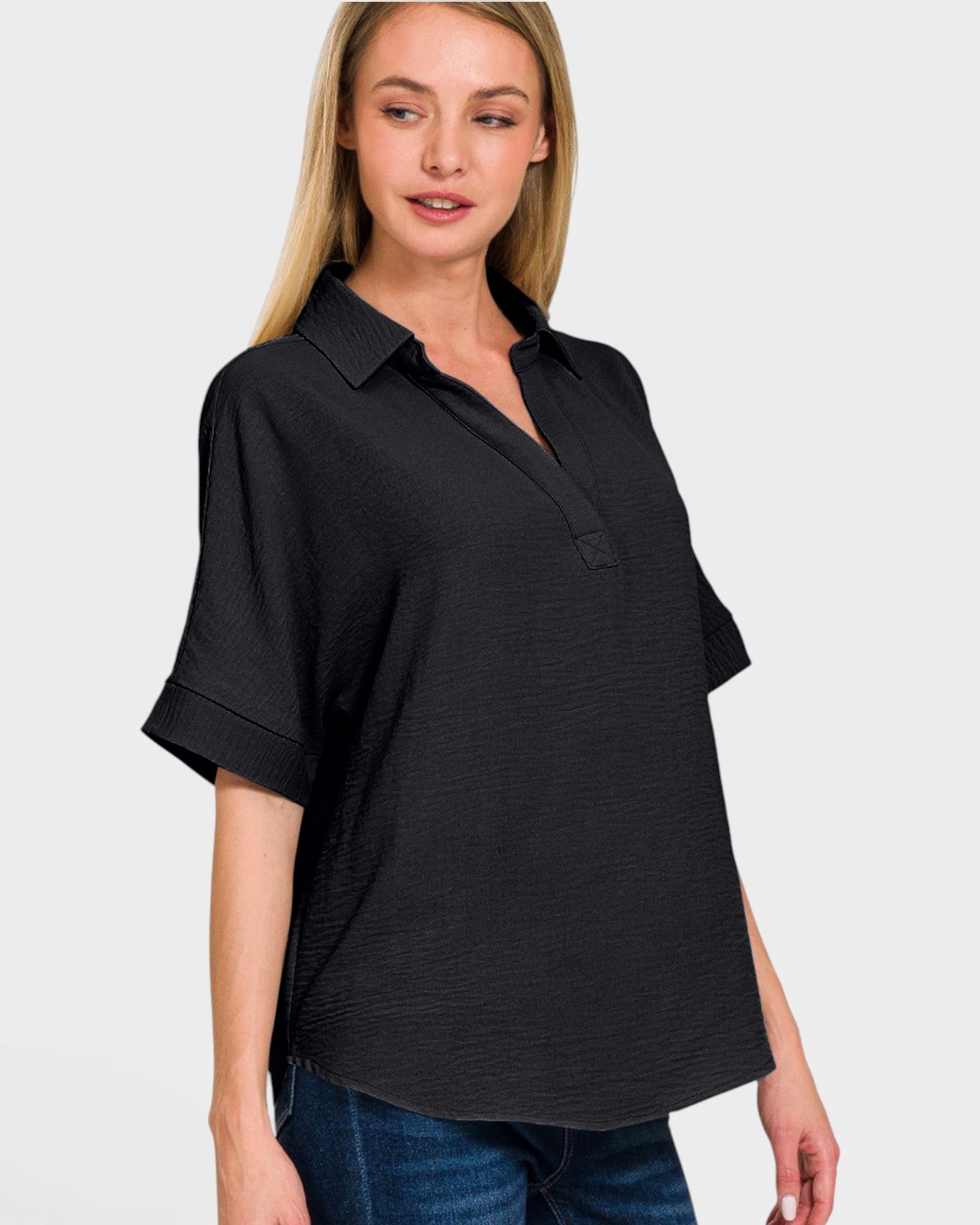 A woman wearing a black short-sleeve collared button-front top with rolled cuffs, paired with blue jeans, photographed against a light gray studio background.