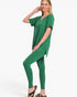 A woman modeling a matching green lounge set consisting of a short-sleeve V-neck top and high-waisted leggings, paired with brown sandals, against a plain white studio background.