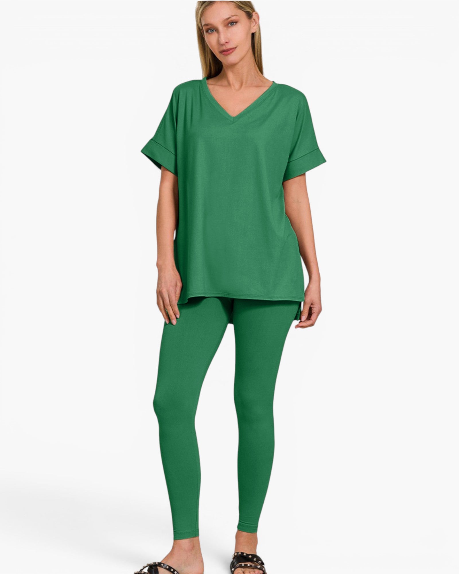 A woman modeling a matching green lounge set consisting of a short-sleeve V-neck top and high-waisted leggings, paired with black studded slide sandals, against a plain white studio background.