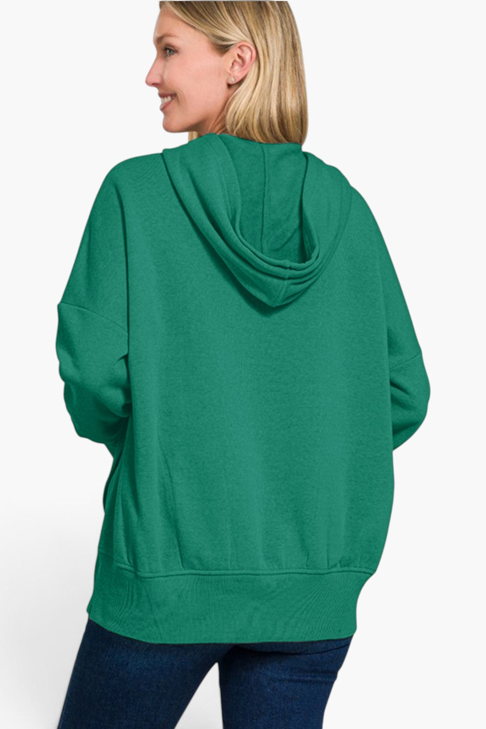 A woman shown from the back wearing an oversized green pullover hoodie with a draped hood and side pockets, paired with dark blue jeans, against a plain white background.