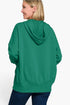 A woman shown from the back wearing an oversized green pullover hoodie with a draped hood and side pockets, paired with dark blue jeans, against a plain white background.