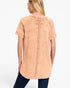 A woman shown from the back wearing a short-sleeve tunic top in a washed peach-beige color, paired with dark blue jeans, against a plain white studio background.