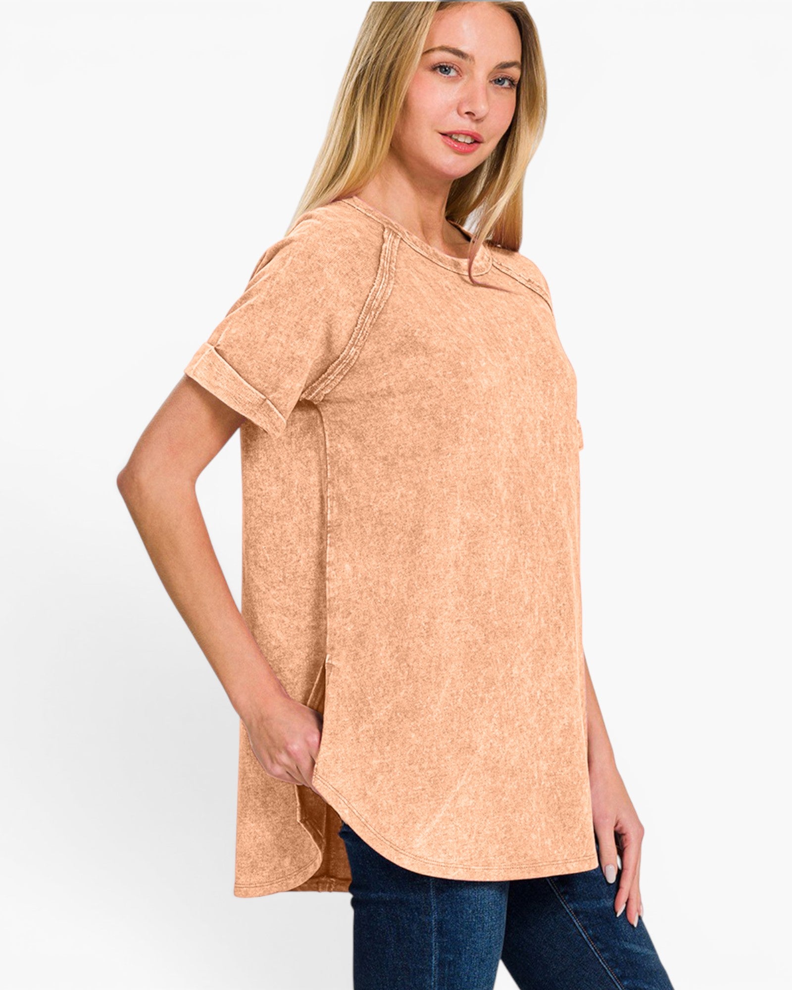 A woman modeling a short-sleeve, crew-neck tunic top in a washed, faded peach-beige color, paired with dark blue jeans against a plain white studio background.
