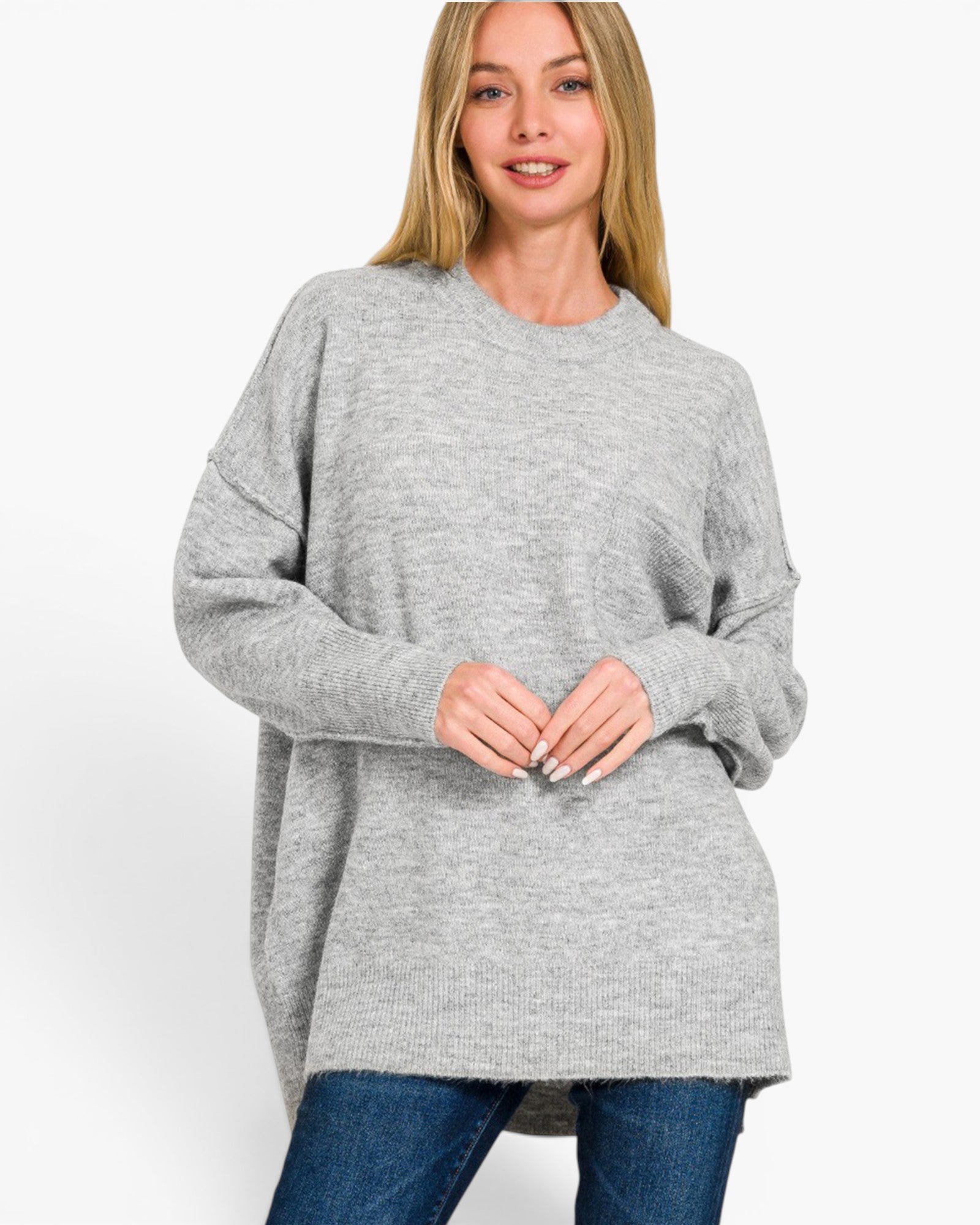 A woman wearing a light gray oversized crewneck sweater and blue jeans, standing against a plain white studio background.