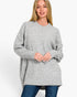 A woman wearing a light gray oversized crewneck sweater and blue jeans, standing against a plain white studio background.