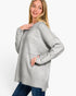 A woman modeling an oversized light gray knit sweater with a large chest pocket, paired with blue jeans, against a plain white studio background.