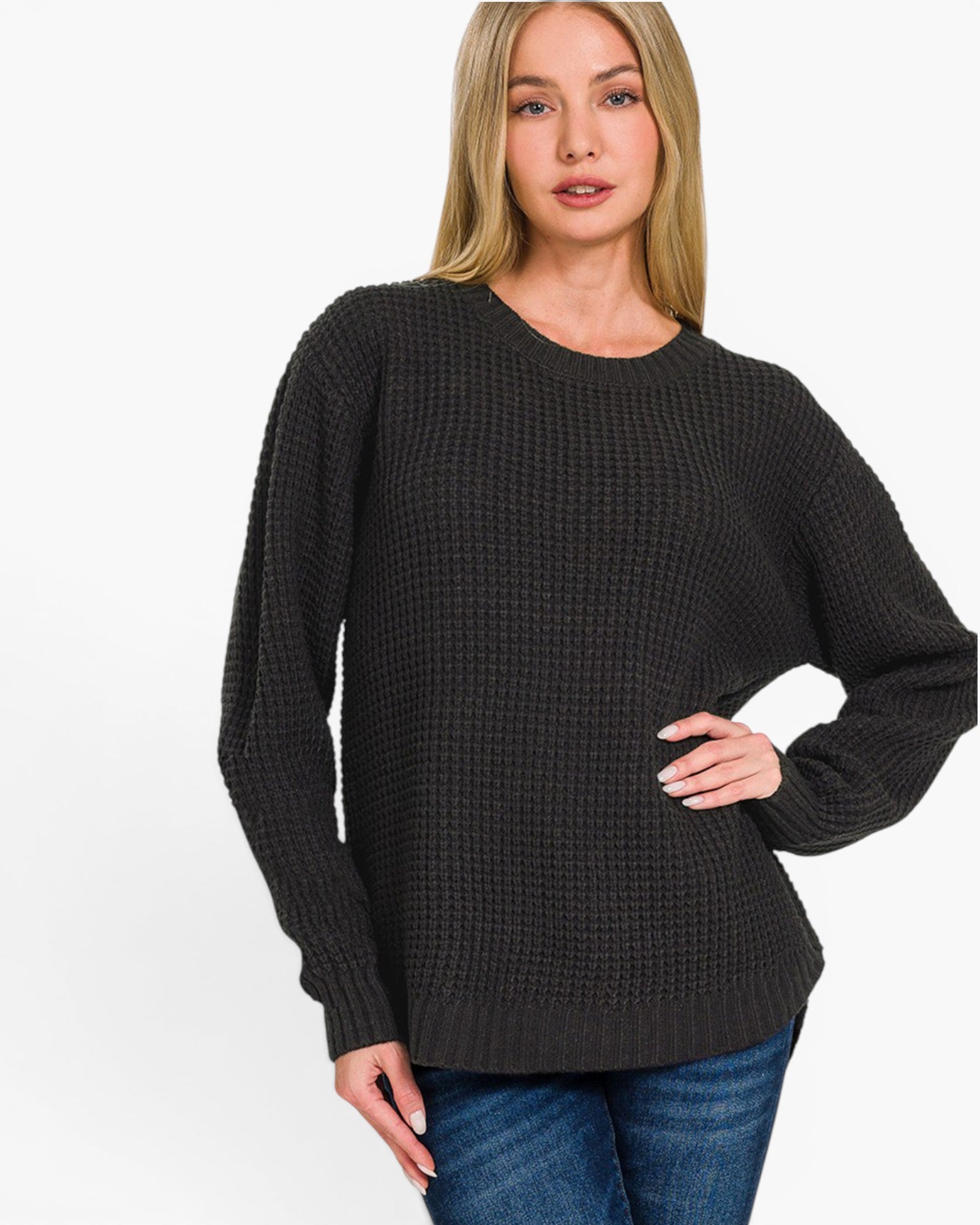 A woman wearing a dark charcoal gray waffle-knit crewneck sweater and blue jeans, standing against a plain white studio background.