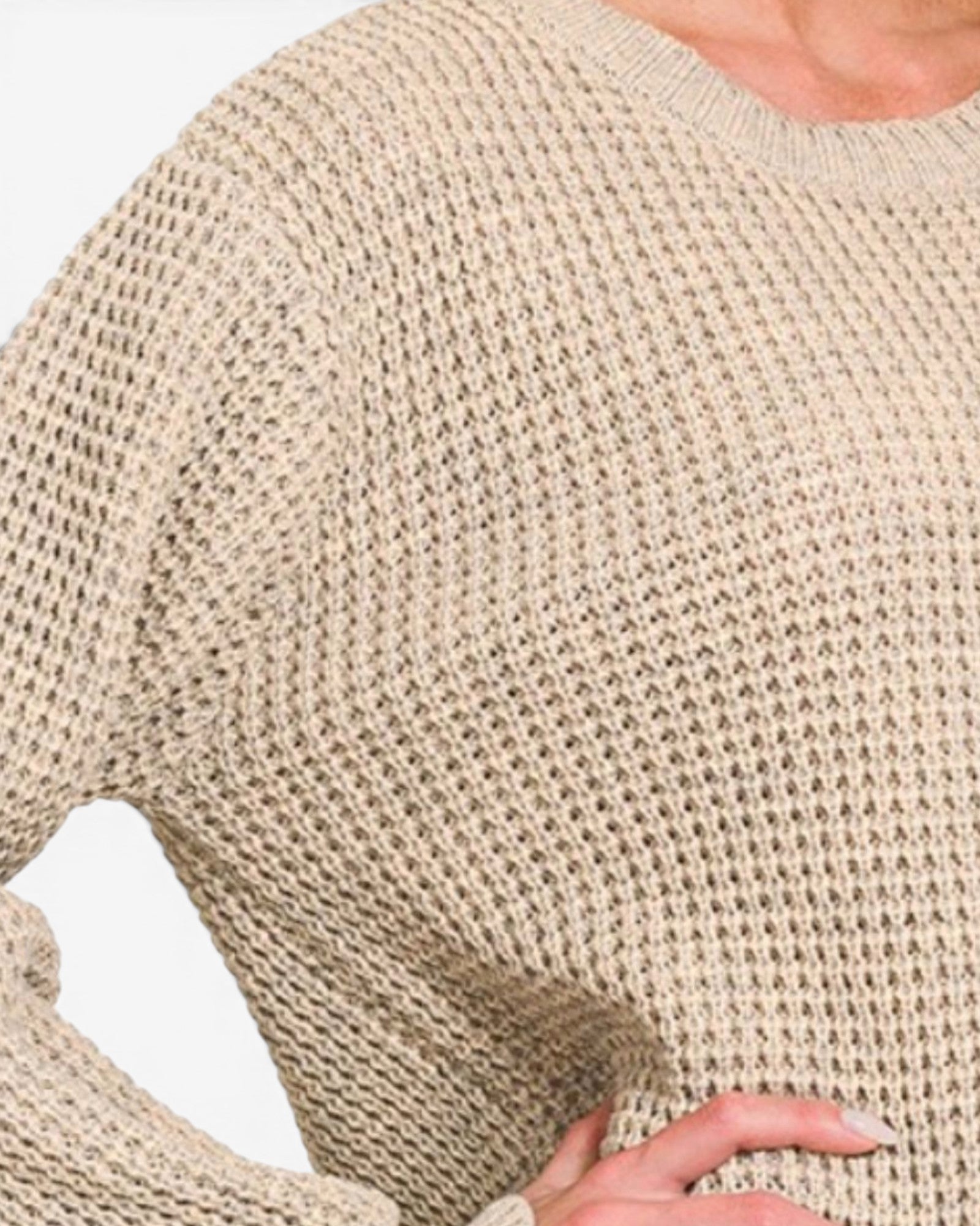 Close-up of a person wearing a chunky knit sweater in a warm beige or oatmeal color, showing the textured open-knit pattern and part of the hand.