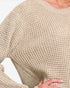 Close-up of a person wearing a chunky knit sweater in a warm beige or oatmeal color, showing the textured open-knit pattern and part of the hand.