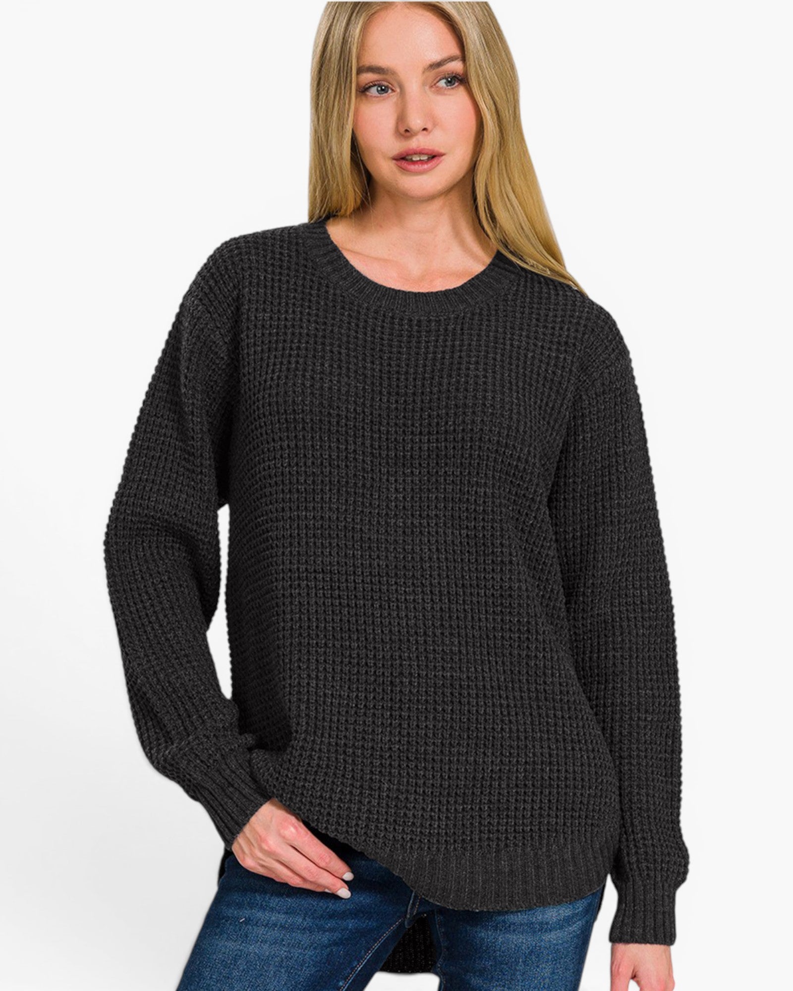 A woman wearing a dark charcoal gray waffle-knit crewneck sweater and blue jeans, photographed against a clean white studio background.