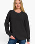 A woman wearing a dark charcoal gray waffle-knit crewneck sweater and blue jeans, photographed against a clean white studio background.