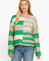 A woman modeling a fuzzy knit sweater with wide green, beige, and off-white horizontal stripes, paired with blue jeans against a plain white background.