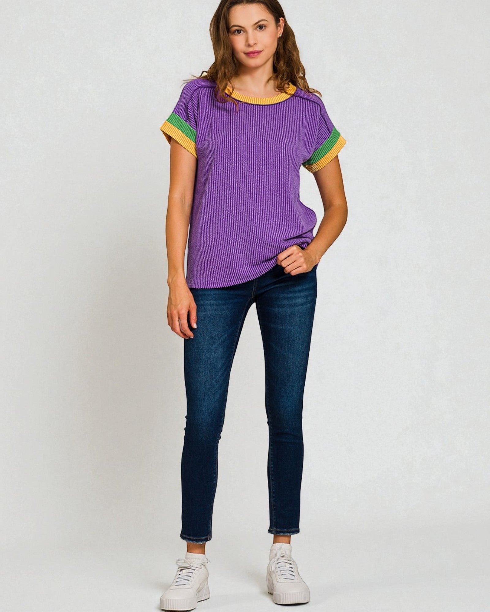 Woman wearing a purple ribbed top with green accents and blue jeans on a white background