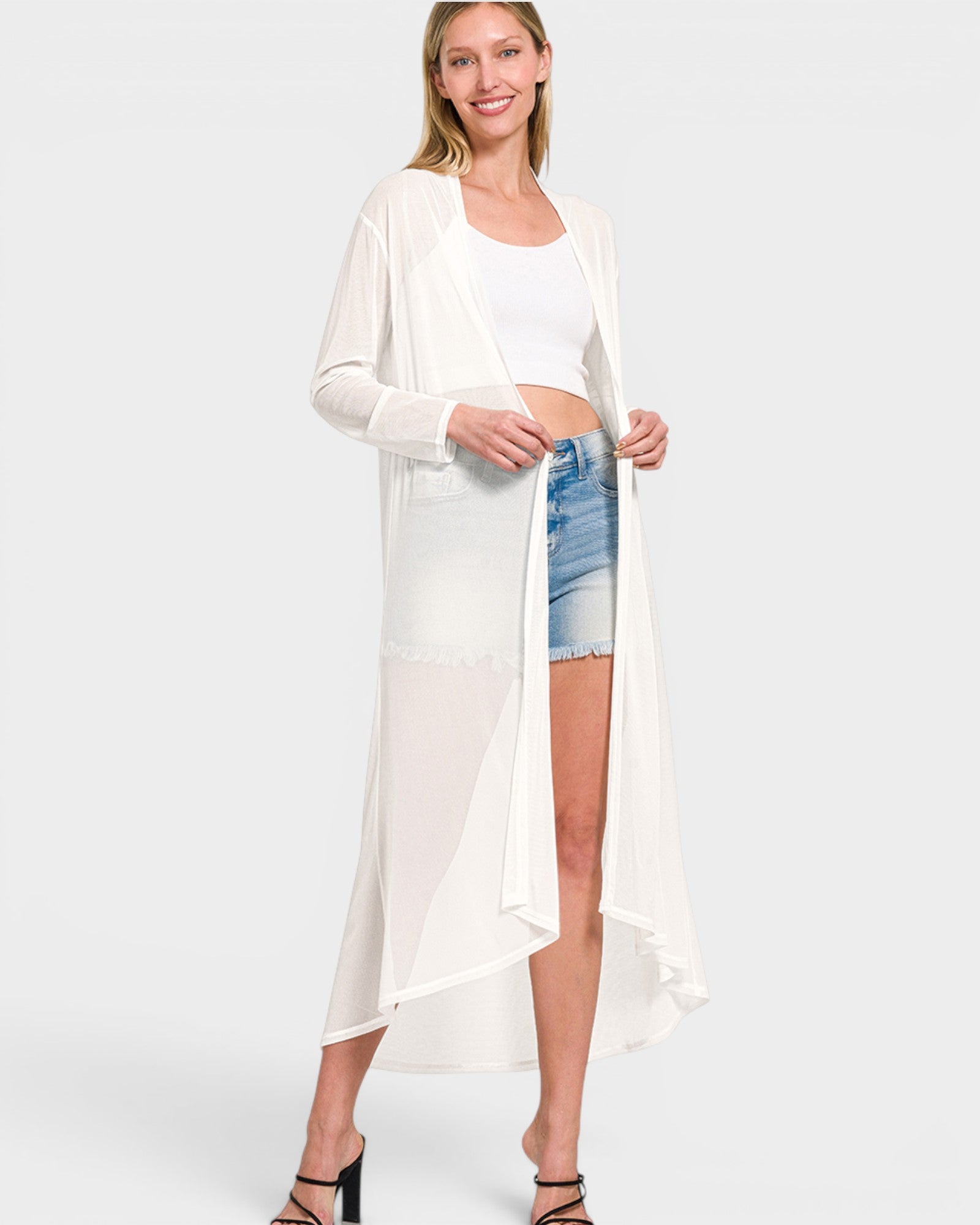 Woman wearing a long white cardigan over a white top and denim shorts on a white background