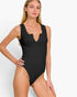 A woman modeling a black sleeveless one-piece swimsuit with a split neckline and ruched detail, photographed against a clean light gray studio background.