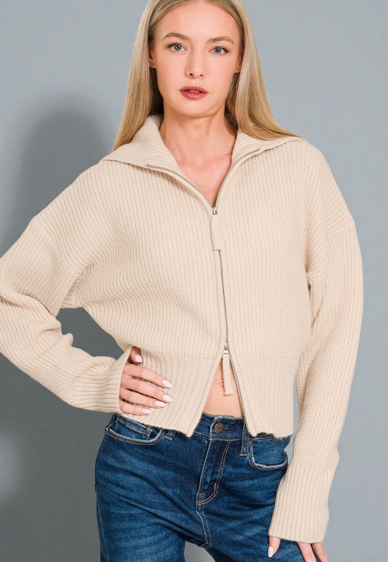 Woman wearing a beige zip-up sweater and blue jeans against a gray background