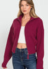 Woman wearing a burgundy zip-up sweater over a white top and blue jeans on a white background