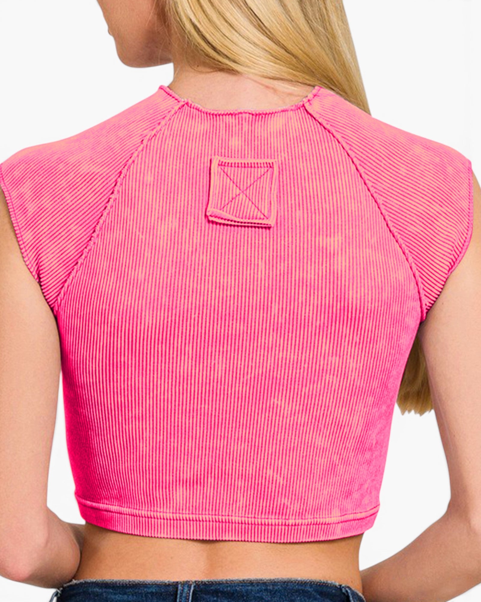 A close-up back view of a woman wearing a bright pink ribbed crop top with raglan sleeves and a small square patch detail on the upper back.