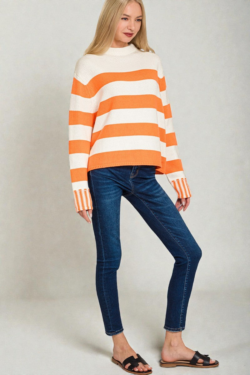 Woman wearing an orange and white striped sweater with blue jeans on a plain background