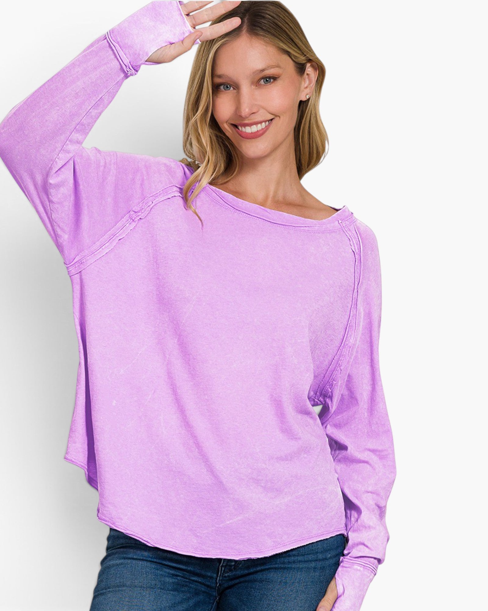A woman wearing a lavender purple long-sleeve top with a wide boat neckline and diagonal seam details, paired with blue jeans, against a plain white background.