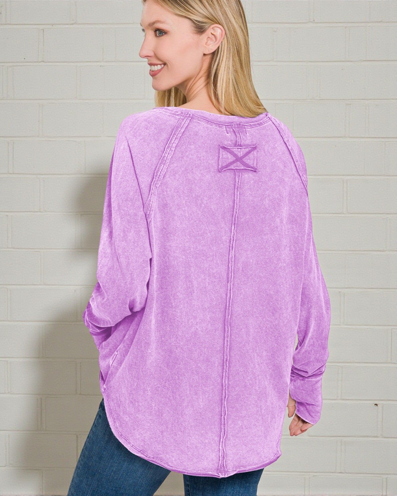 Woman wearing a purple long-sleeve shirt with a logo on the back against a light gray brick wall.