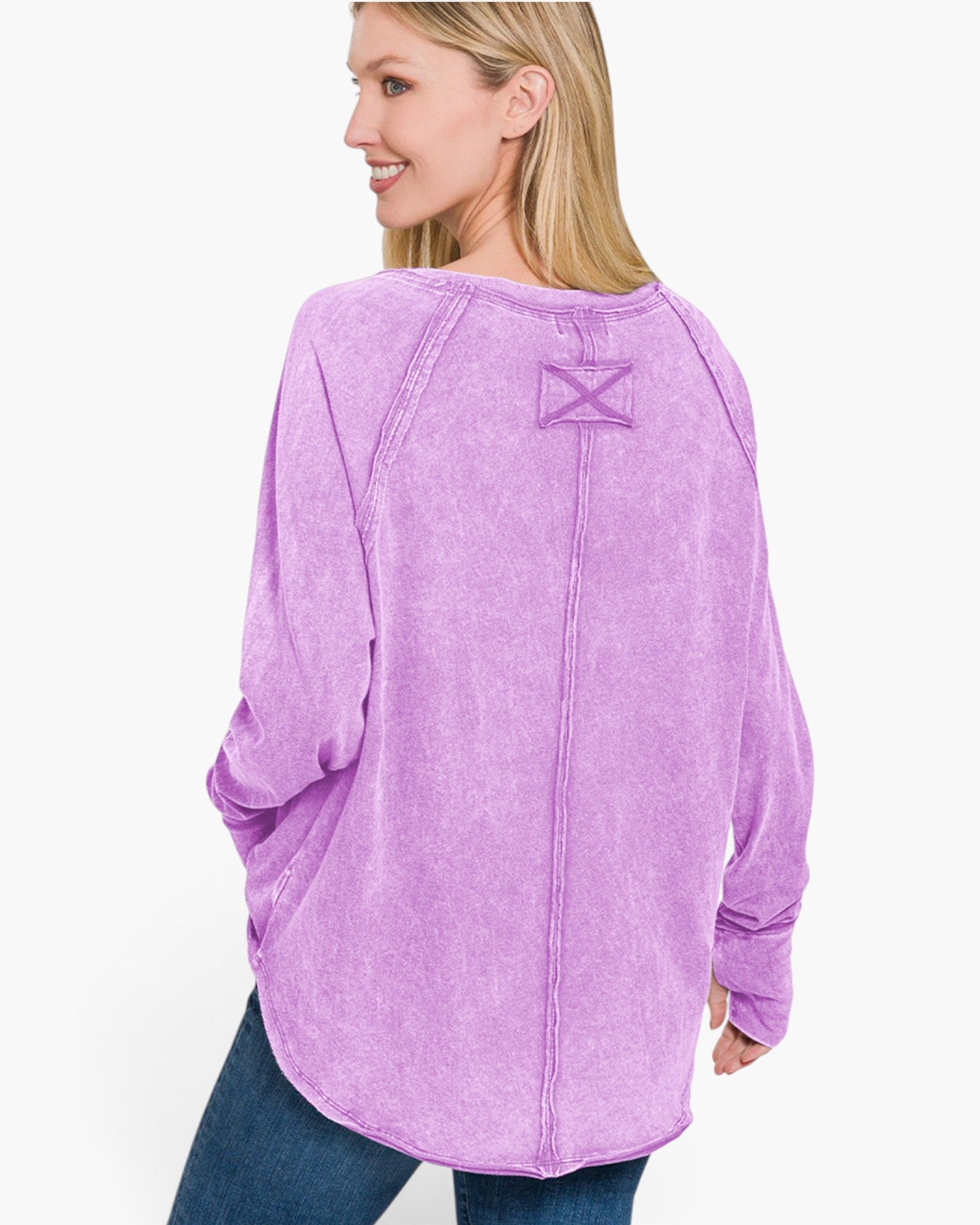 A woman wearing a lavender purple long-sleeve top with a unique crisscross lace-up detail on the back neckline, paired with blue jeans, shown from a three-quarters back view against a white studio background.
