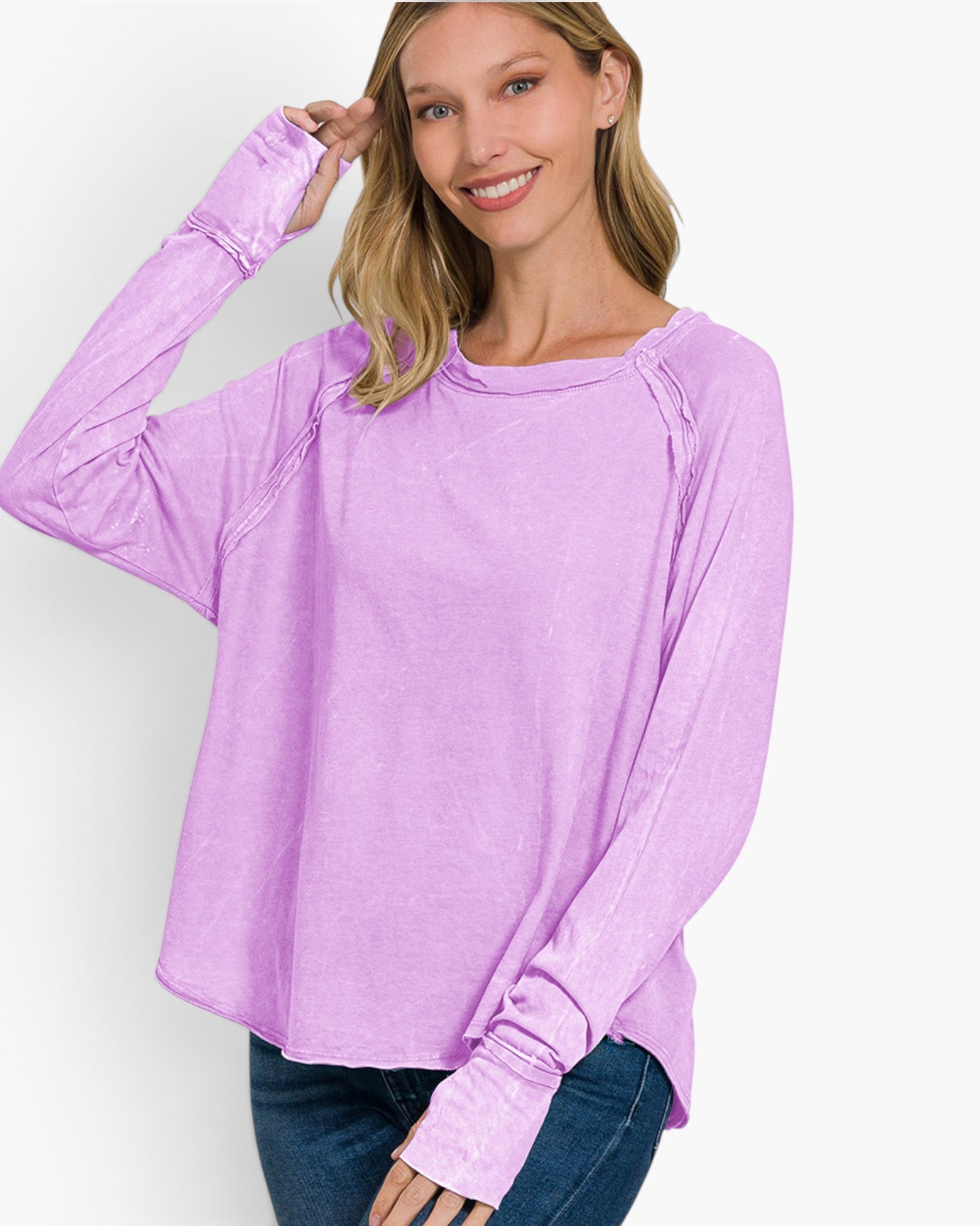 A woman wearing a lavender long-sleeve top with ruched details on the sleeves and a wide scoop neckline, paired with dark blue jeans, against a plain white background.
