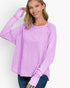 A woman wearing a lavender long-sleeve top with ruched details on the sleeves and a wide scoop neckline, paired with dark blue jeans, against a plain white background.