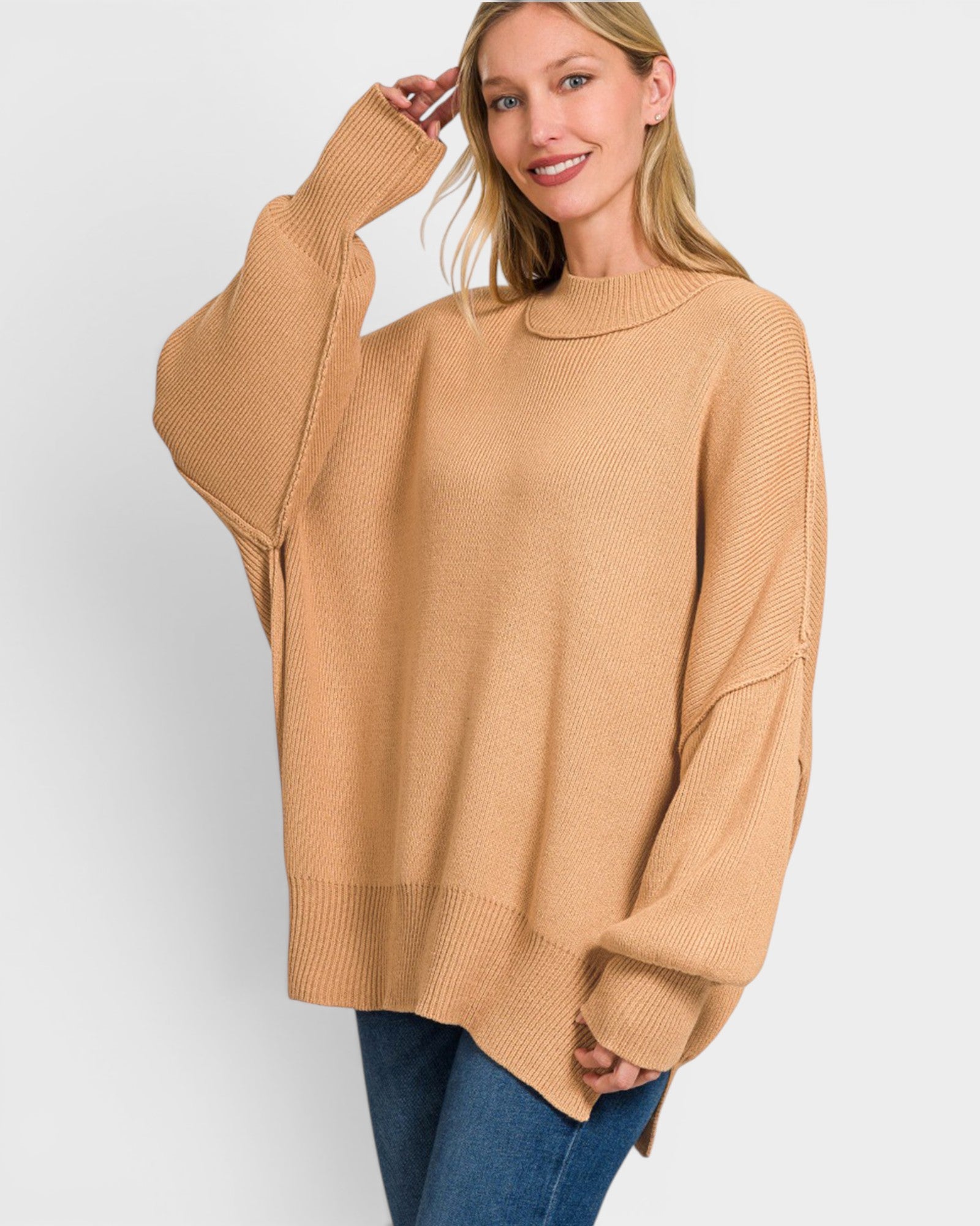 A woman wearing a camel-colored knit sweater with wide balloon sleeves and a crew neck, paired with blue jeans, against a light gray studio background.
