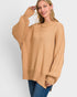 A woman wearing a camel-colored knit sweater with wide balloon sleeves and a crew neck, paired with blue jeans, against a light gray studio background.