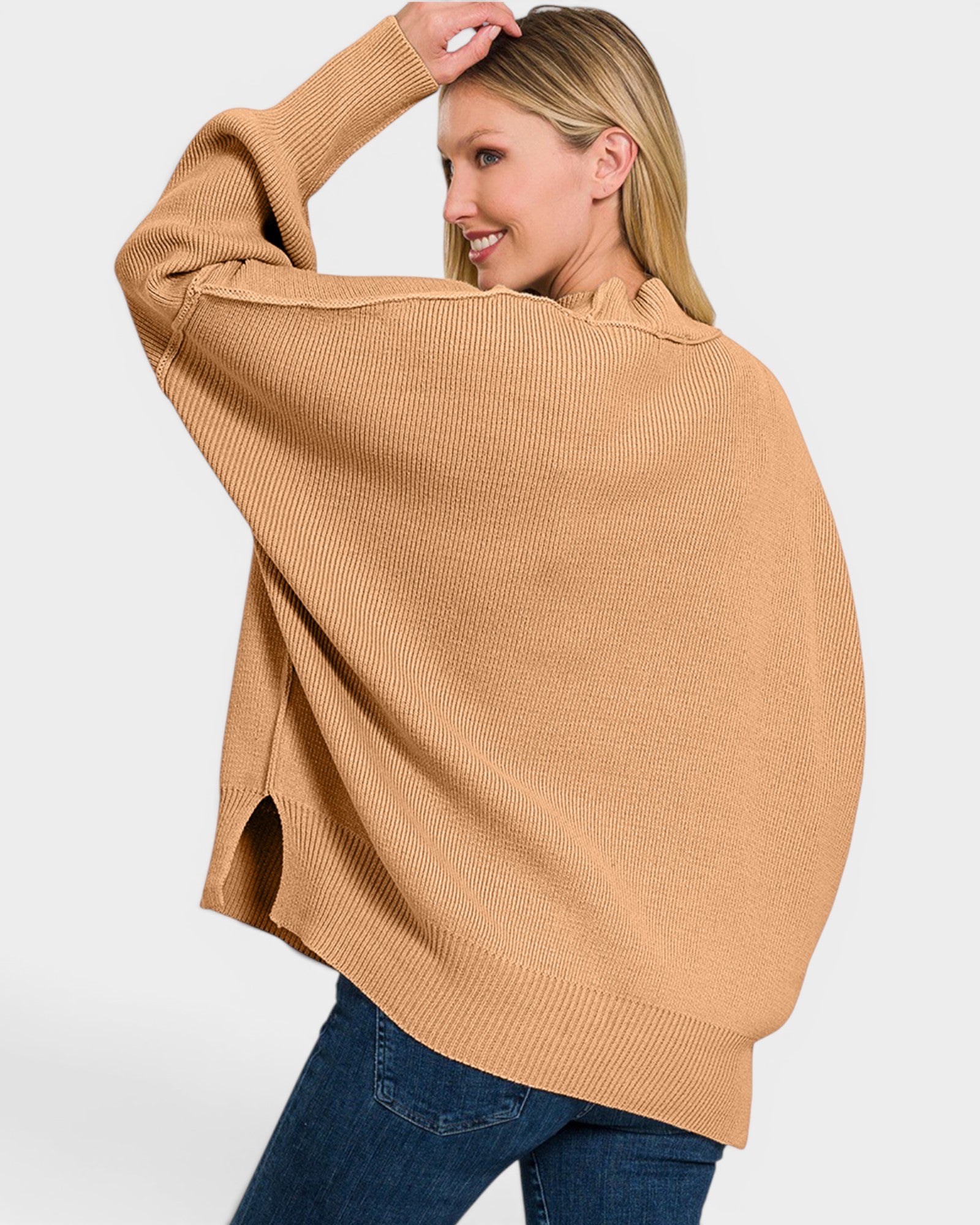 A woman wearing a camel-colored ribbed knit sweater with a relaxed, oversized fit and blue jeans, photographed against a light gray studio background.