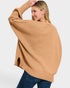 A woman wearing a camel-colored ribbed knit sweater with a relaxed, oversized fit and blue jeans, photographed against a light gray studio background.