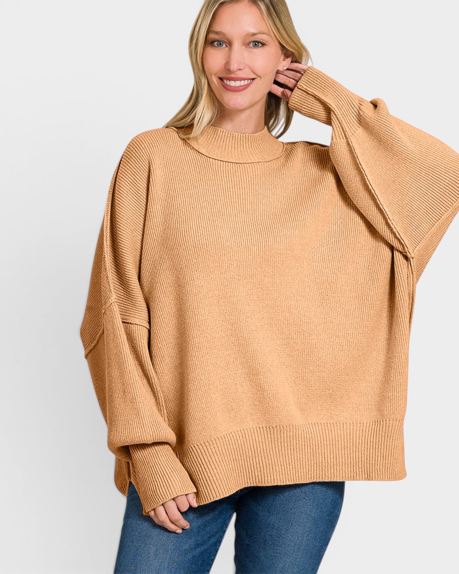 A woman wearing a camel-colored ribbed knit sweater with balloon sleeves and blue jeans, photographed against a light gray studio background.