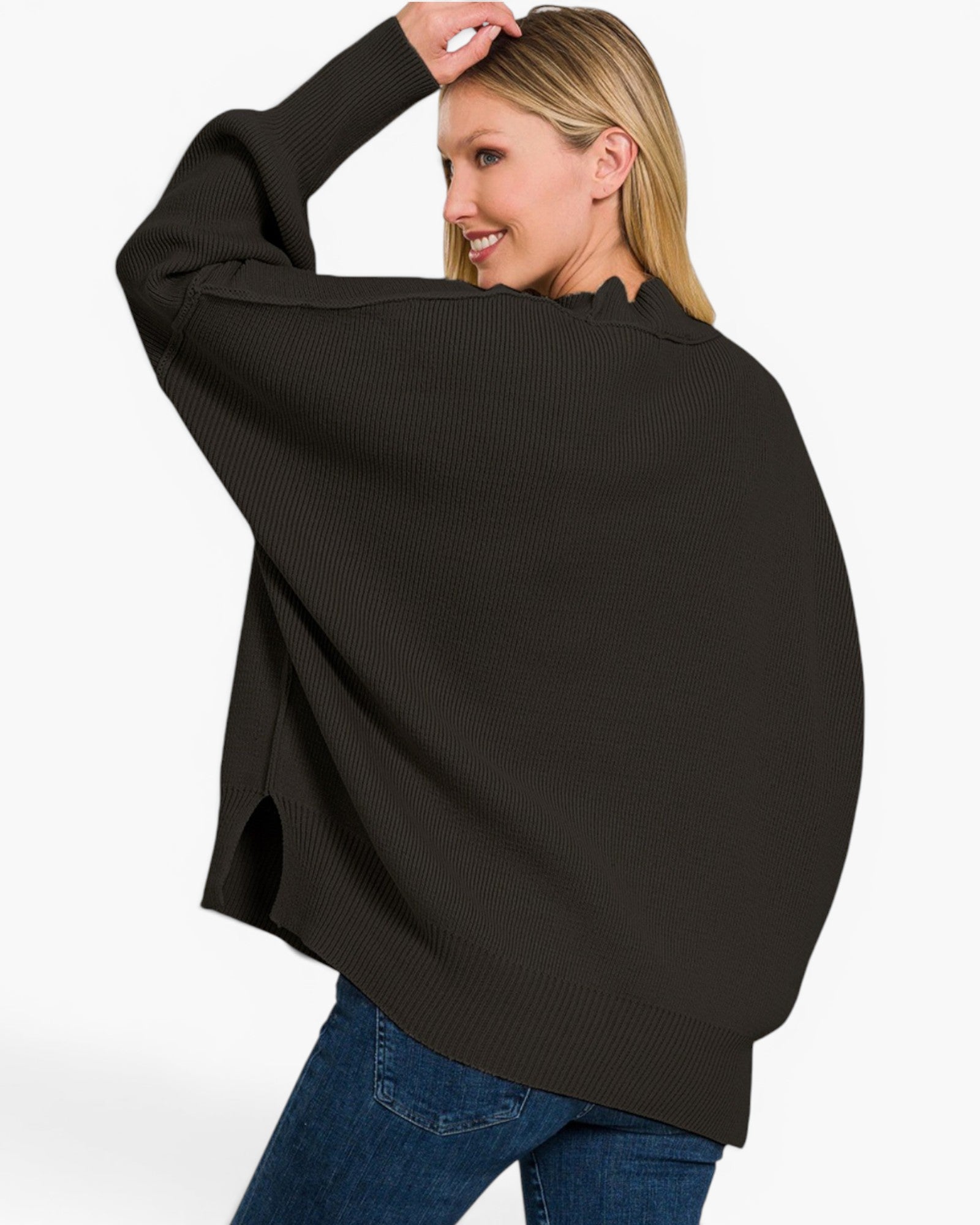 A woman wearing a black ribbed knit poncho-style sweater with blue jeans, shown from the back against a plain light gray studio background.