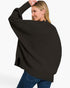 A woman wearing a black ribbed knit poncho-style sweater with blue jeans, shown from the back against a plain light gray studio background.