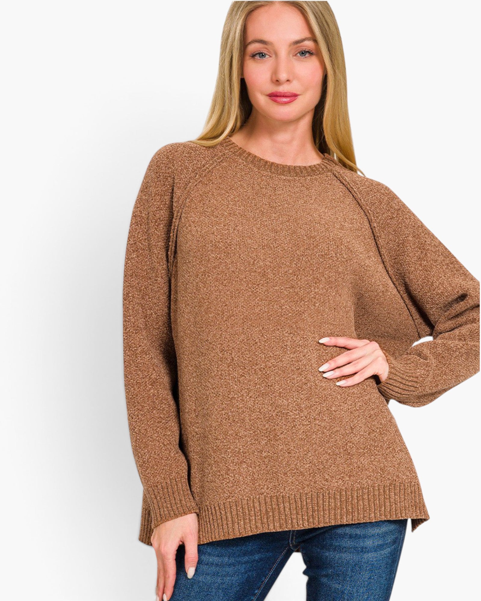 A woman wearing a brown knit sweater with blue jeans, standing against a plain white studio background.