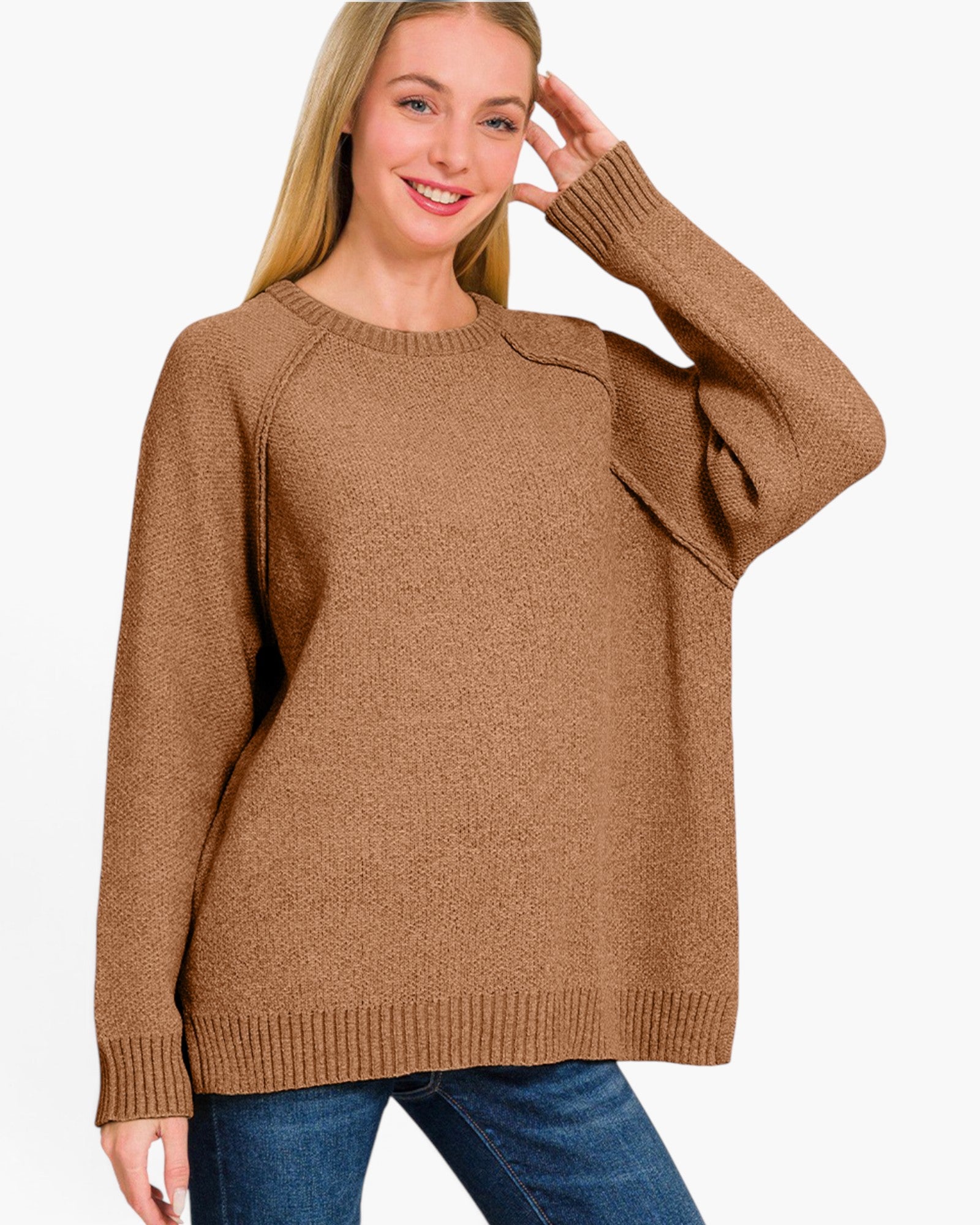 A woman wearing a brown knit crewneck sweater with raglan sleeves and a draped pocket detail on the upper sleeve, paired with blue jeans against a plain white background.